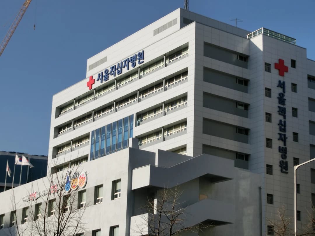 A multi-story hospital building with signage and a red cross emblem seen from the street