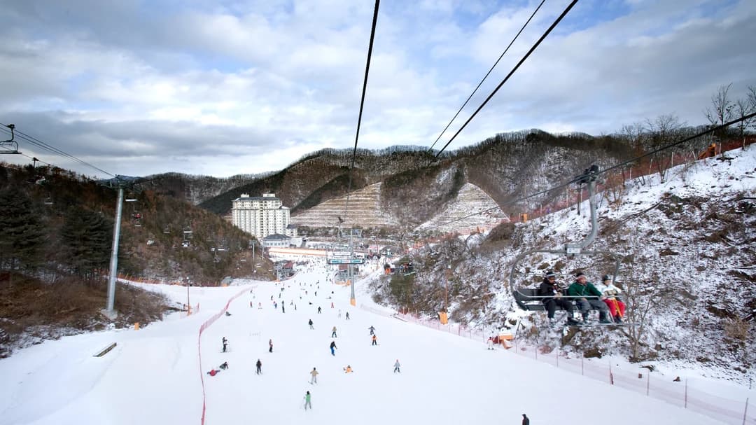 View down a groomed ski slope from a chairlift, with skiers below and the resort hotel at the base (Elysian Gangchon)